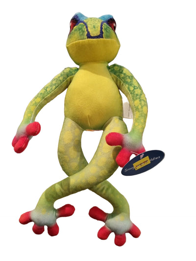 A plush frog toy standing upright on a glass display shelf. The frog is bright yellow-green with darker green mottled spots, blue and red facial details, and red-pink feet and hands.