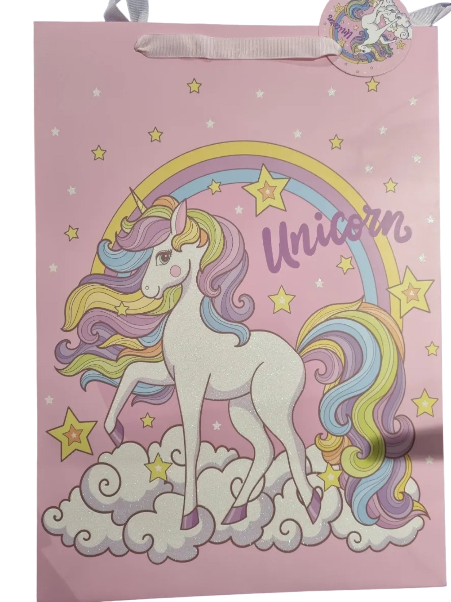 A pink unicorn-themed party decoration or backdrop featuring a white unicorn with a pastel rainbow mane, yellow stars, and the word 'Unicorn' in purple script. A matching round tag with a ribbon is attached on the left side.